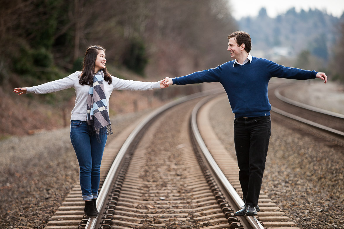 Seattle wedding photographer Tom Ellis Photography. Couple holds hands while balancing on railroad tracks during their engagement photo session near Golden Gardens Park in Seattle